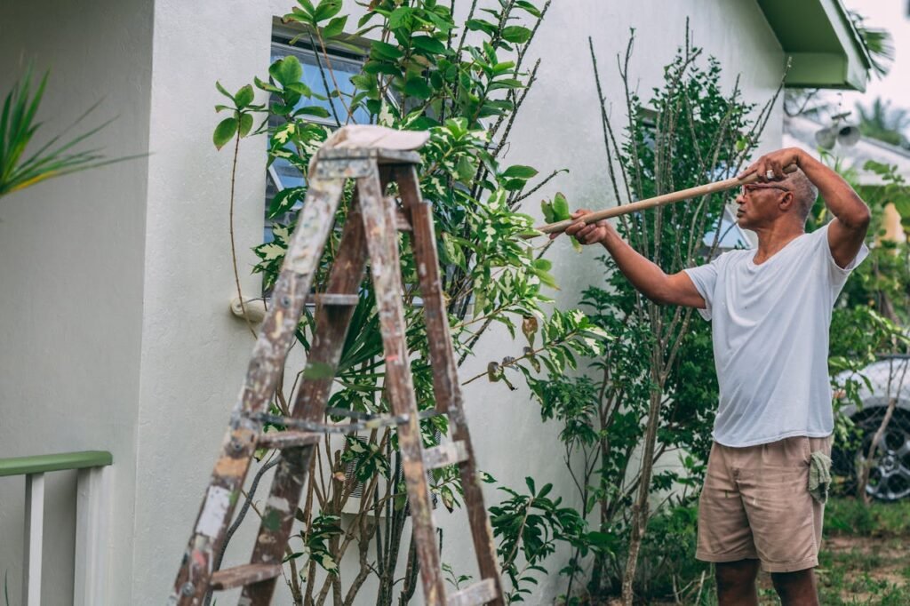 An adult man paints a house wall outdoors with a roller and ladder in a garden.