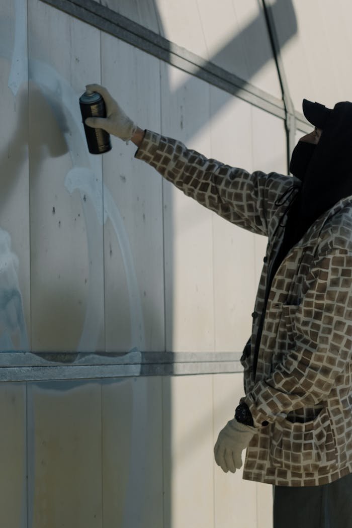 A masked artist in gloves painting graffiti on an outdoor wall with spray paint.